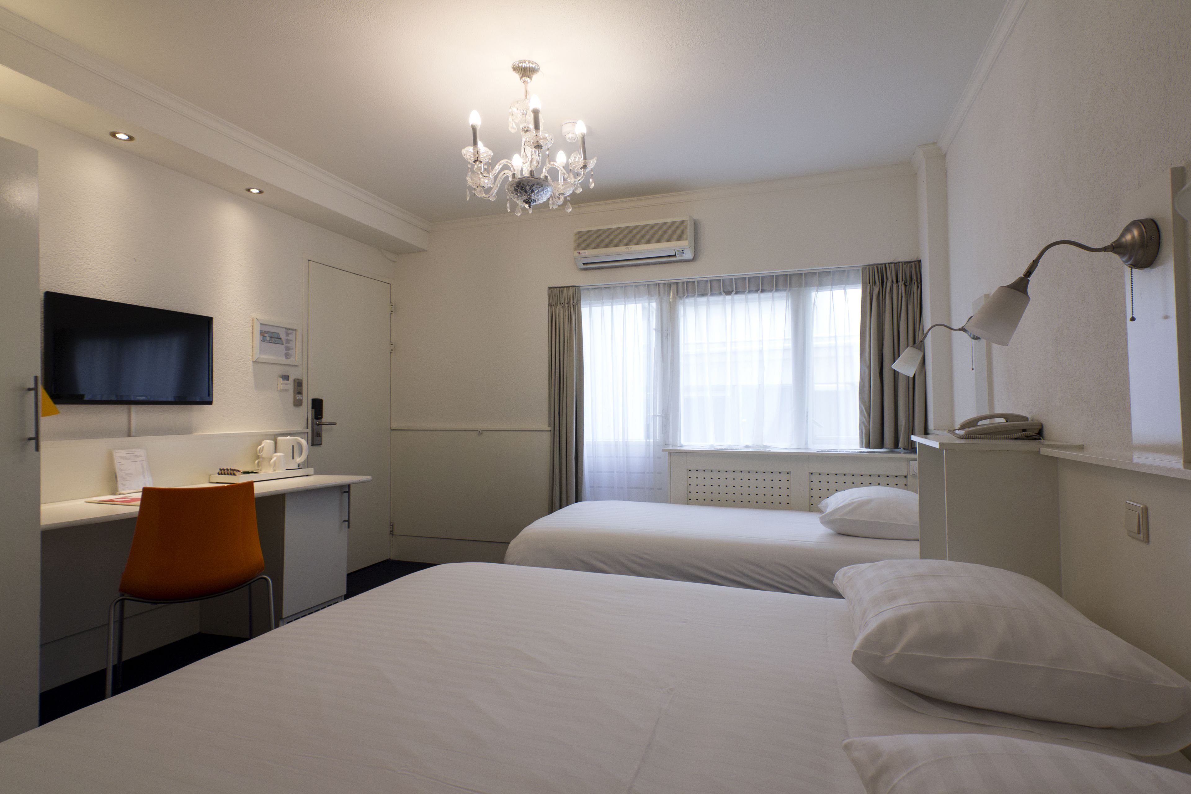 A hotel room with two beds, a desk, a chair, a TV, a chandelier, and a window with curtains.