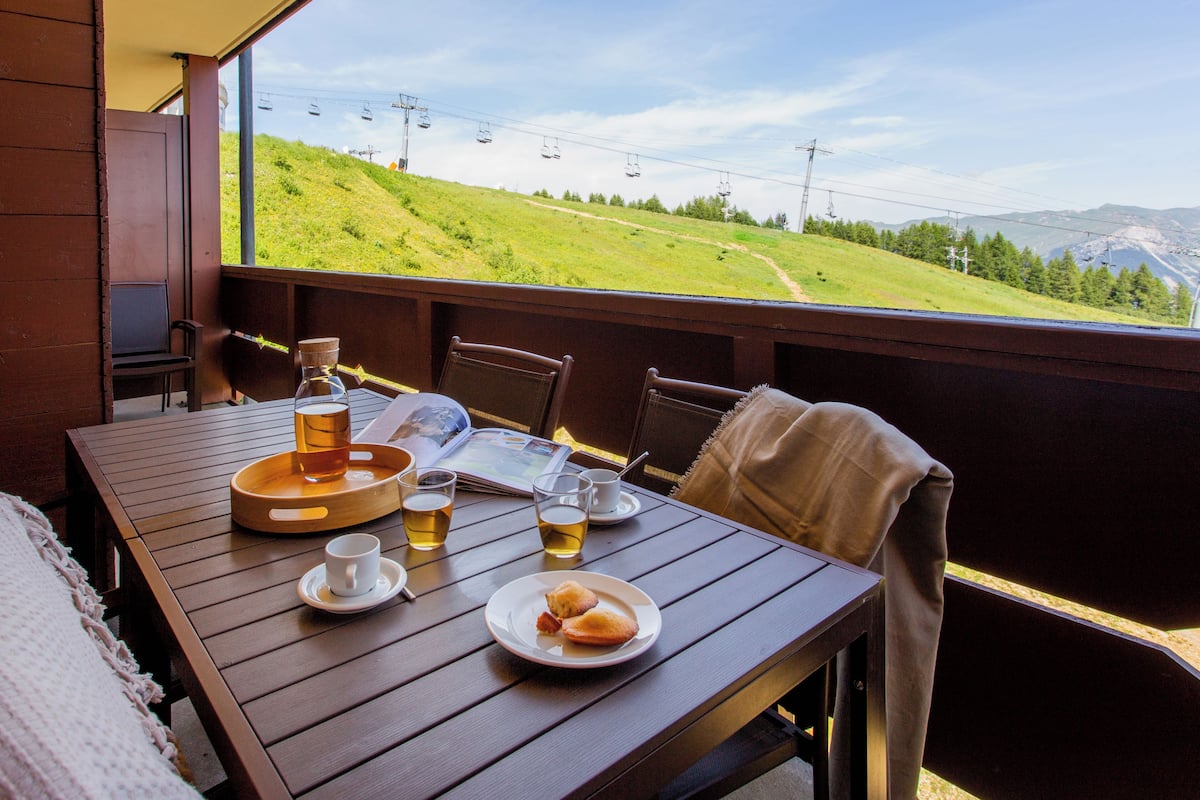 Une table en bois, sur laquelle sont disposés un plateau, des assiettes, des tasses et une bouteille, le tout placé devant un paysage montagneux pittoresque, avec une chaise et une couverture à proximité.