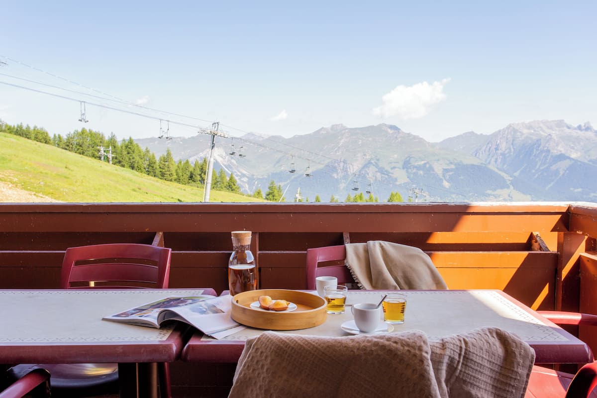 Une table avec un magazine, un bol en bois rempli de viennoiseries, un verre de bière et une bouteille d’eau, le tout placé devant un paysage de montagne avec un télésiège en arrière-plan.