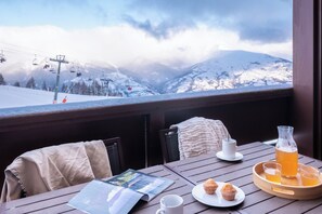 A wooden table with a menu, a glass of orange juice, a muffin, a cup, and a bottle, with a snowy mountain view in the background. - Résidence Pierre & Vacances Premium Les Hauts Bois (Aime-la-Plagne)