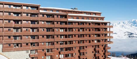 A multi-story hotel with a modern architectural design, featuring balconies and a ski resort backdrop.