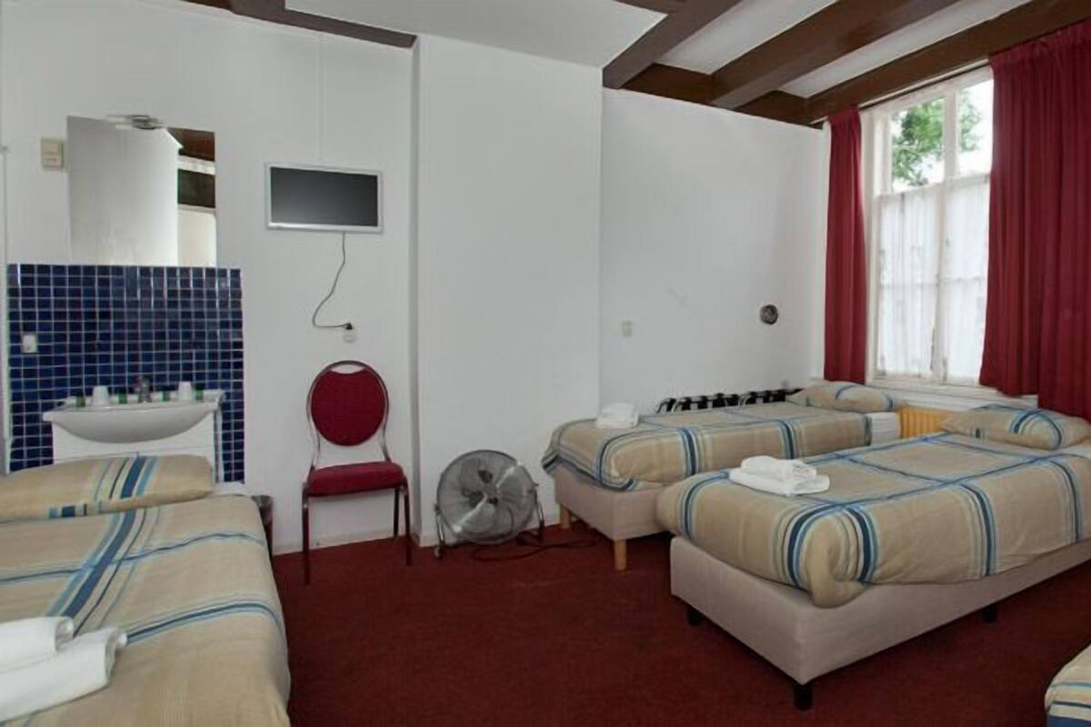 A hotel room with two beds, a red chair, a small table with a white sink, and a window with red curtains.