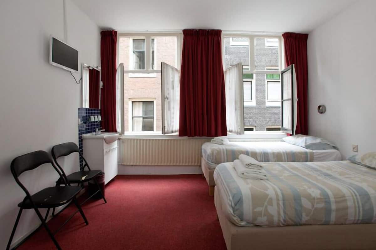 A hotel room with two beds, a desk, a chair, and a window with red curtains.
