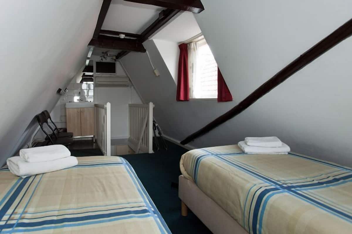 A bedroom with two beds, a small kitchenette, a window with red curtains, and a sloped ceiling.