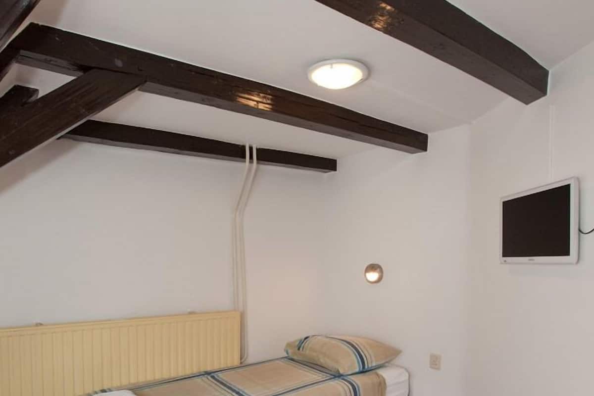 A single bed with a striped bedspread and a white pillow, a small bedside table, a wall-mounted TV, and a ceiling light.