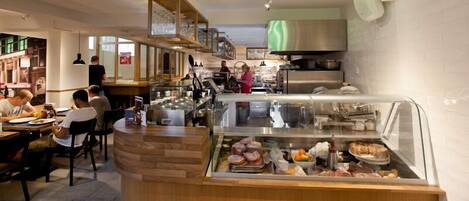 Un restaurante con un mostrador que exhibe diferentes alimentos, una zona de cocina con electrodomésticos de acero inoxidable y clientes comiendo adentro.