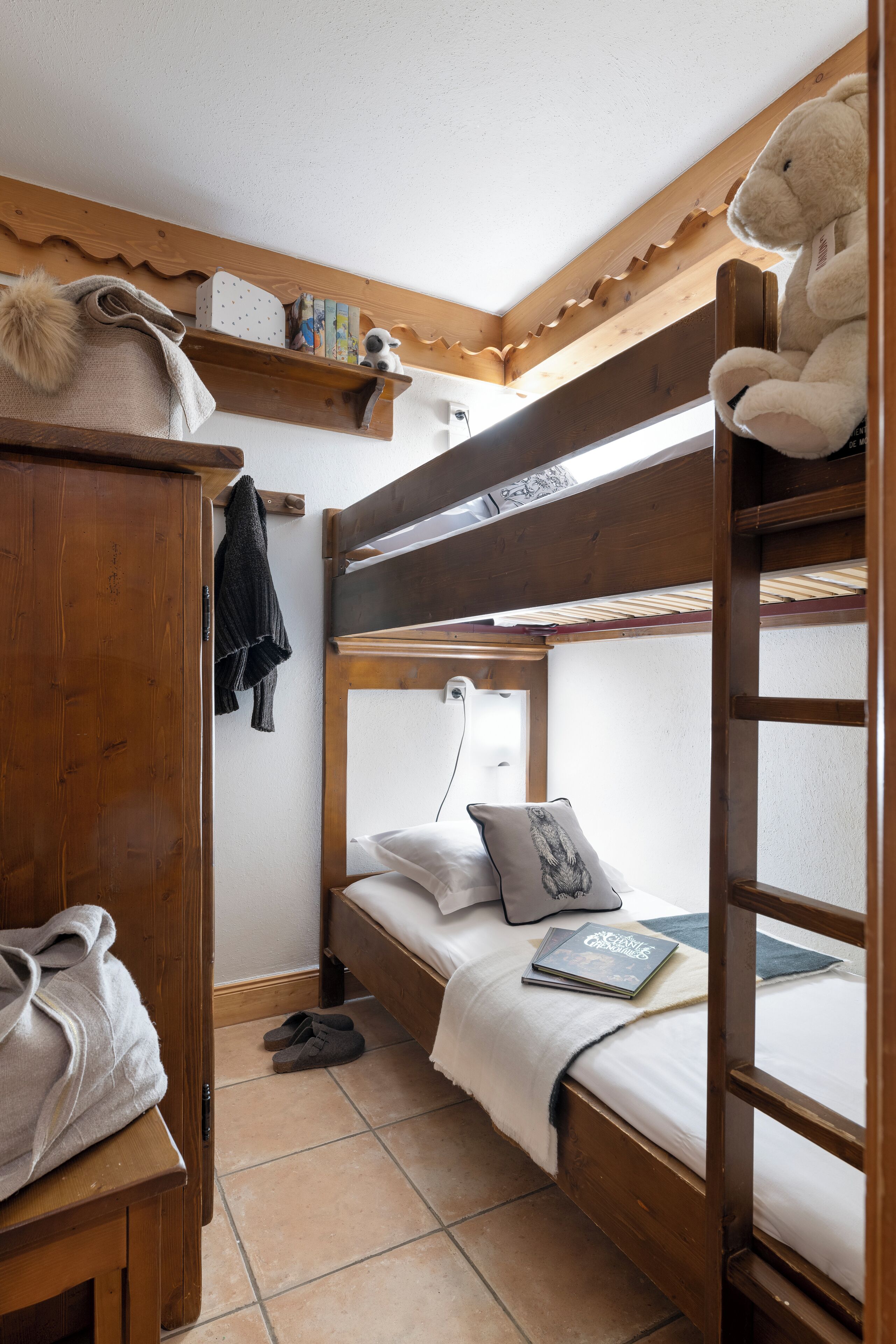 A bunk bed room with a wooden headboard, a shelf with books and a stuffed toy, and a pair of slippers on the floor.