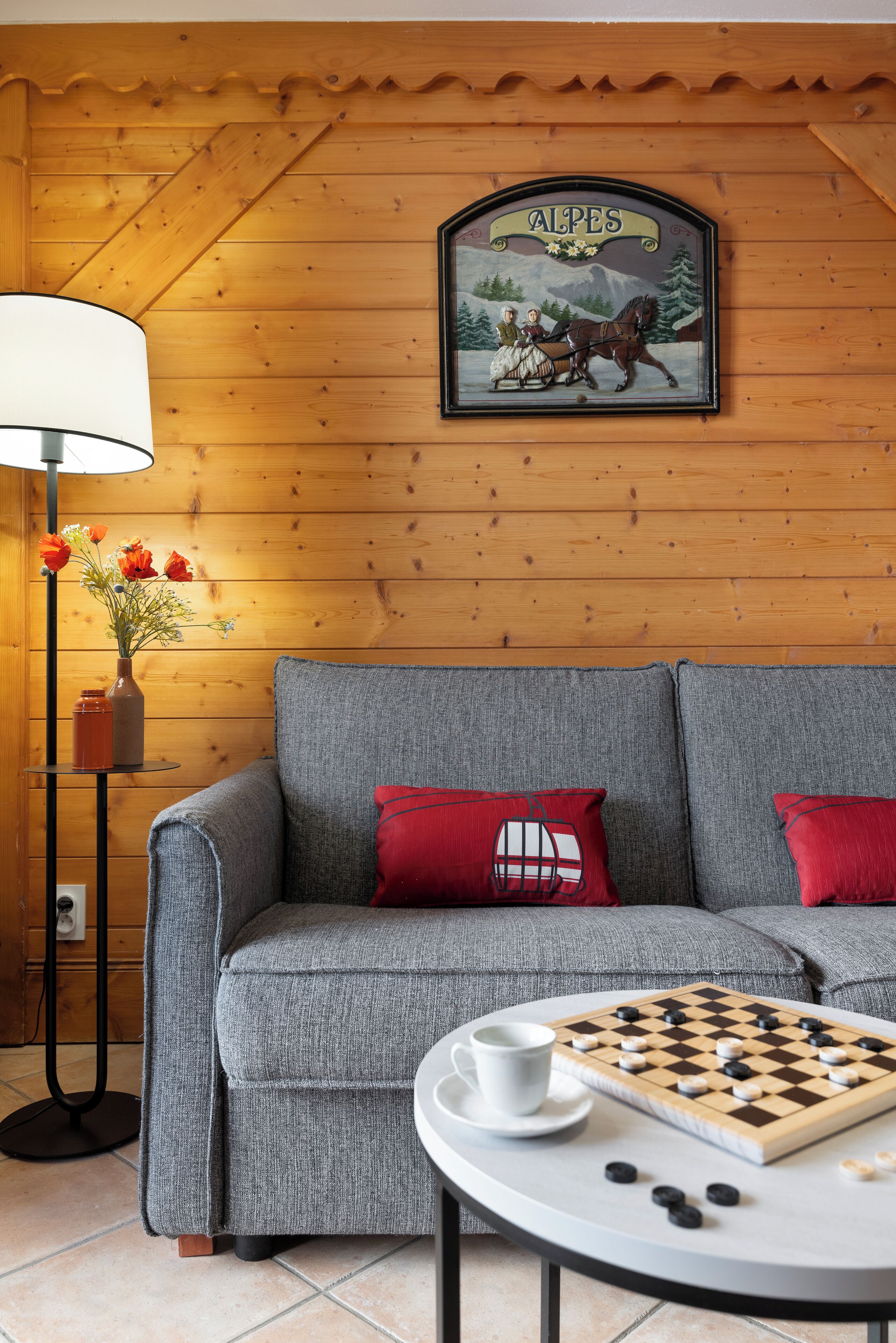 A cozy room with a wooden wall, a grey sofa with red cushions, a chessboard, and a lamp with flowers.