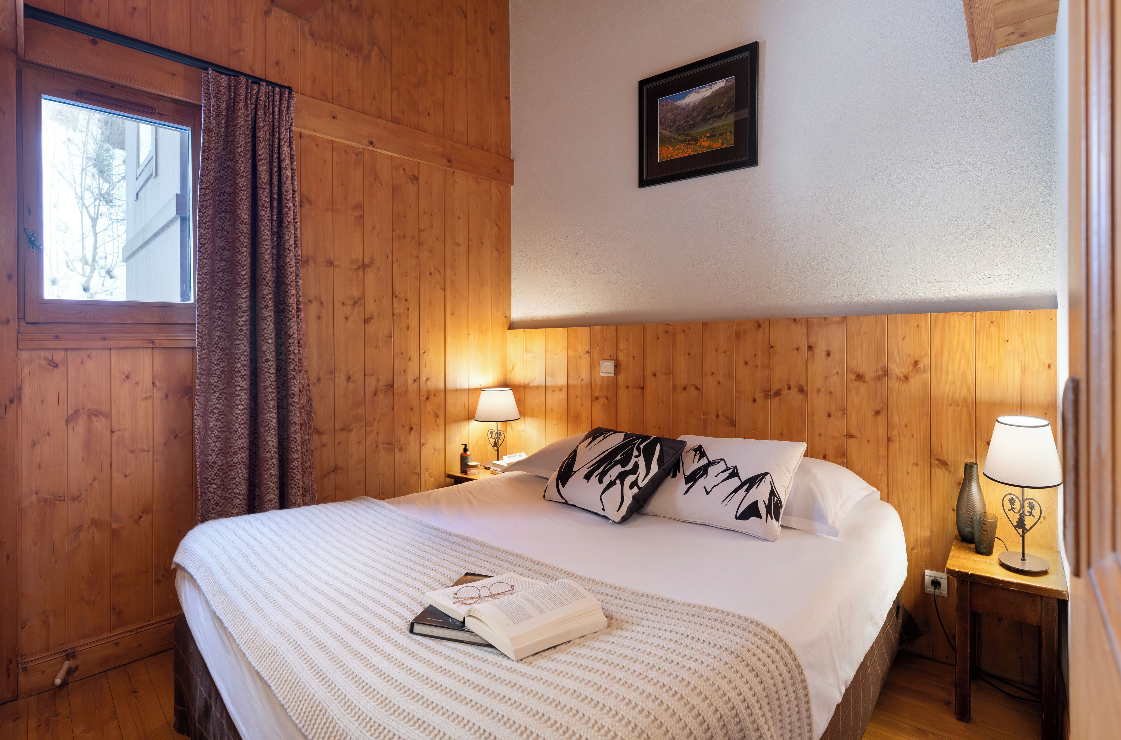Een kamer met houten lambrisering, een bed, twee bedlampen, een ingelijste foto aan de muur en een raam met gordijnen.