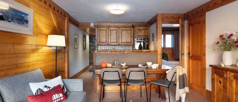 A cozy living room with a grey sofa, a dining table with two chairs, and a kitchen area with wooden cabinets and a built-in oven.