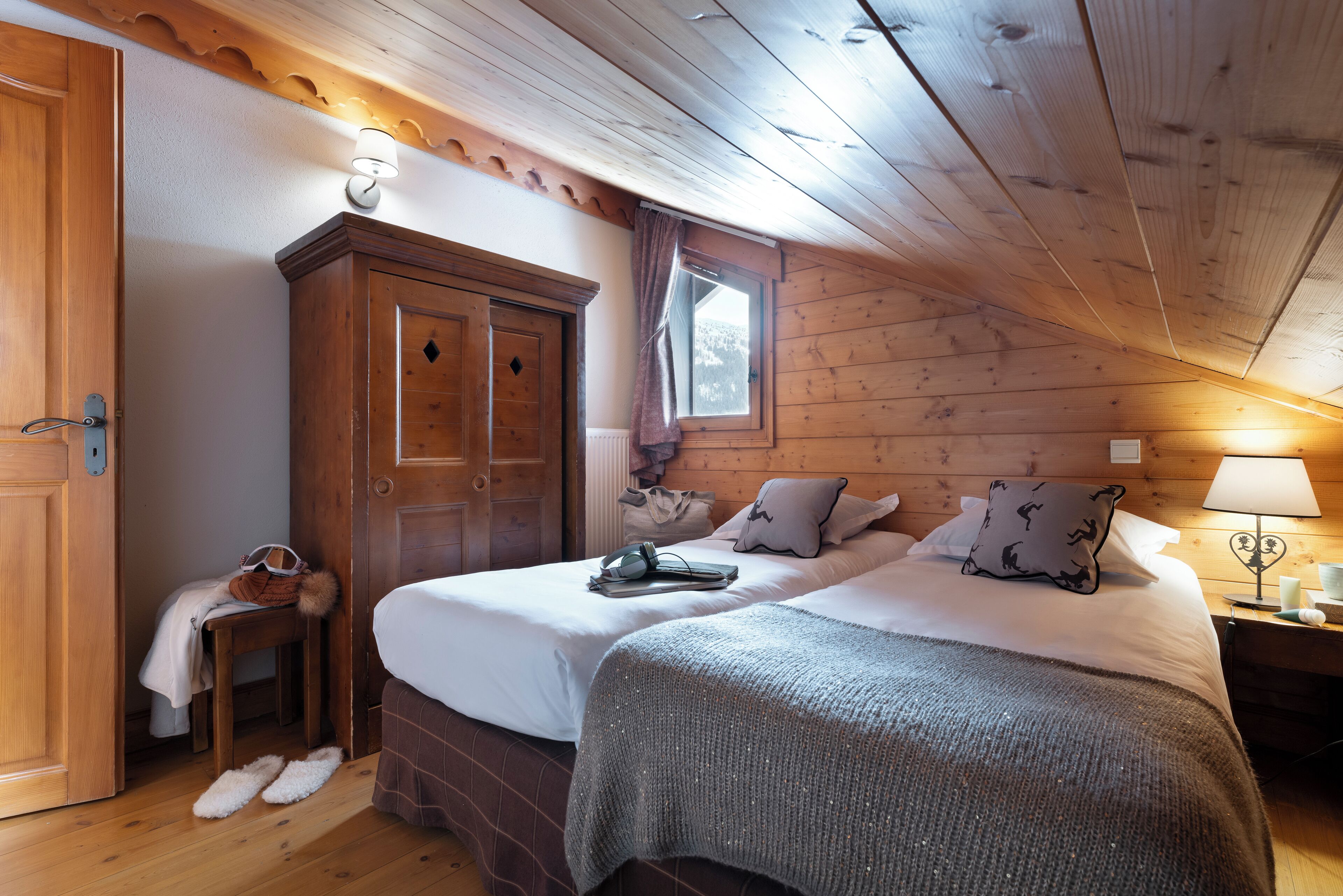 A cozy bedroom with two beds, wooden walls, a window with curtains, a nightstand with a lamp, and a wardrobe.