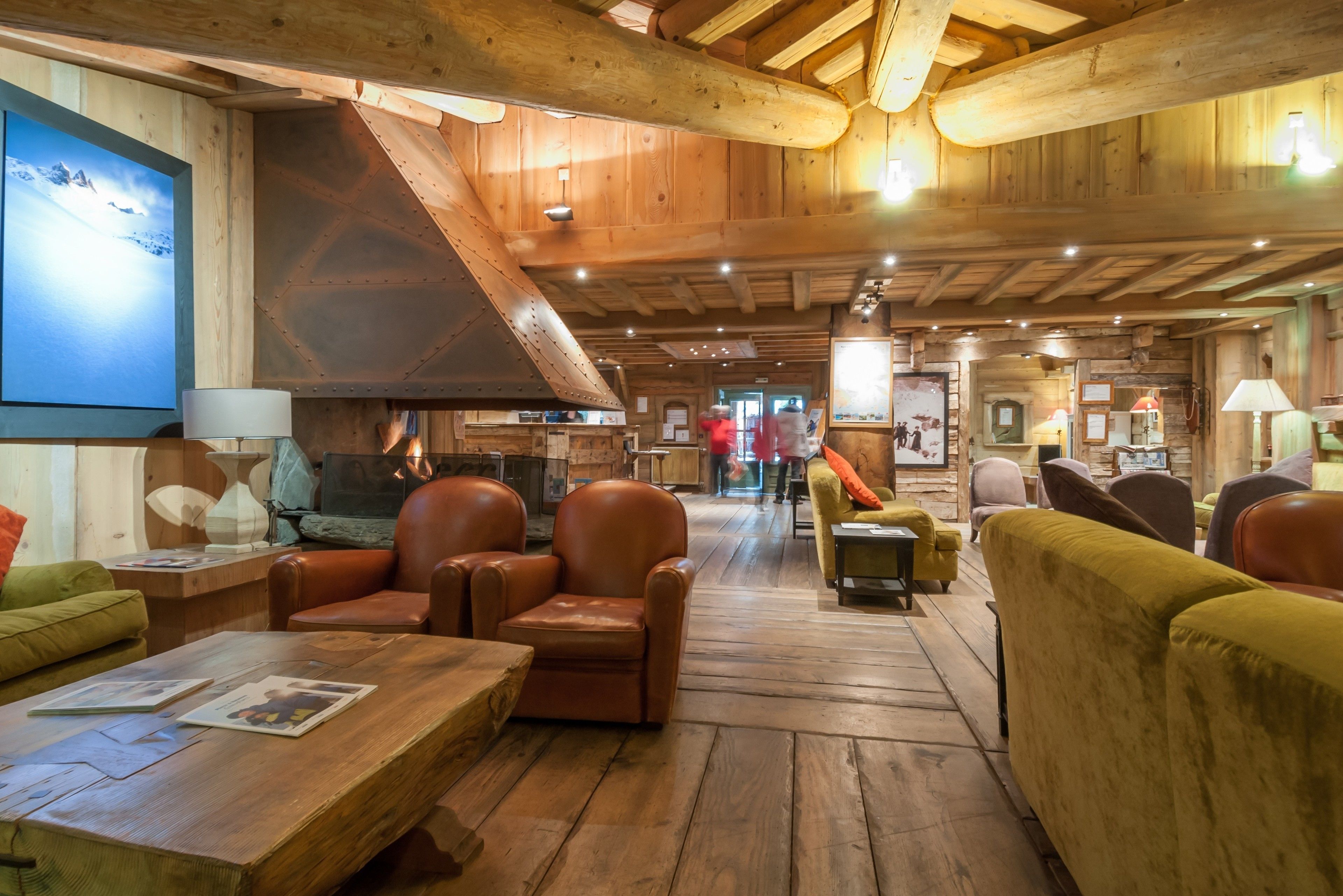 A spacious lobby with wooden floors and high ceilings, featuring leather sofas, a fireplace, and a large TV screen.