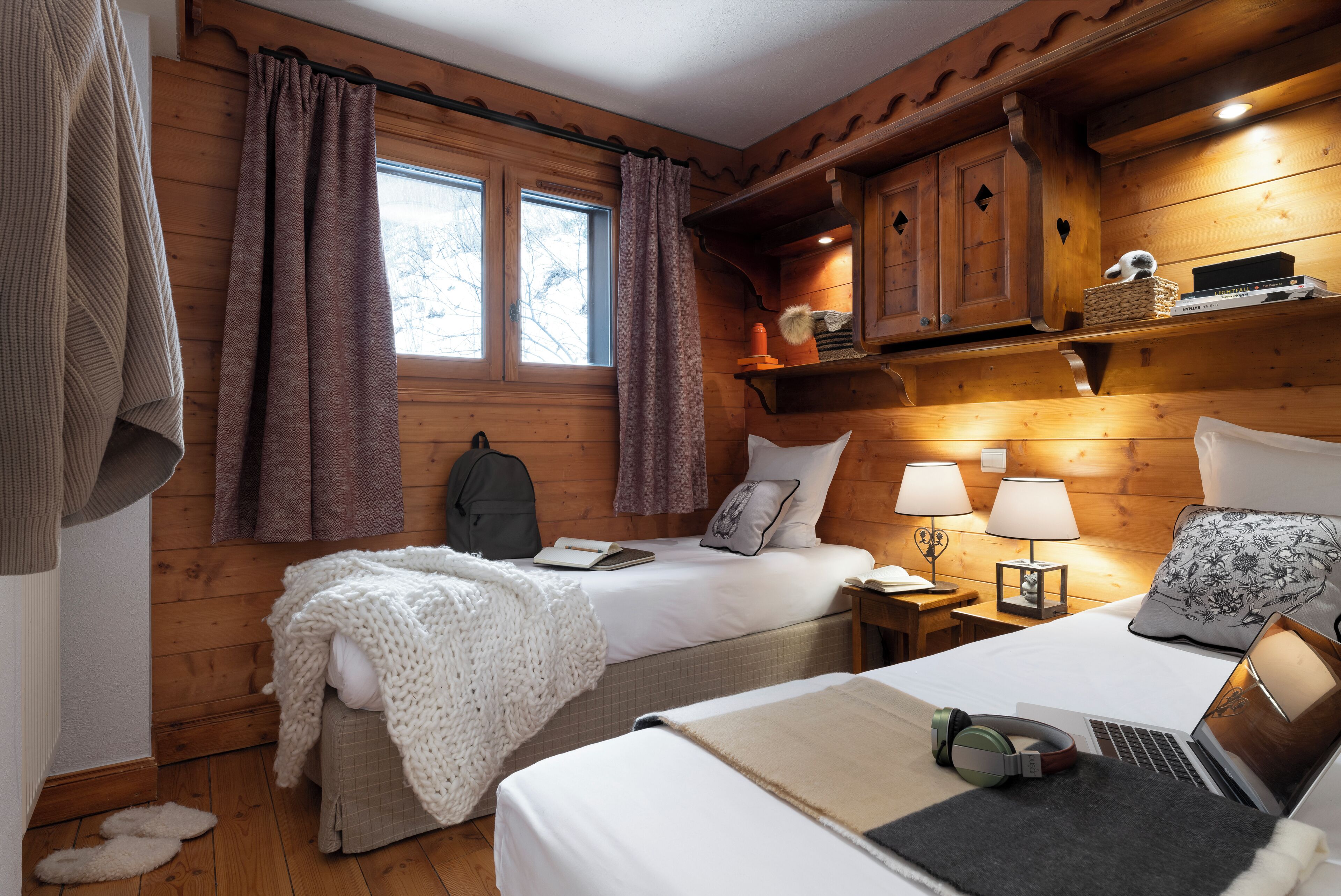 A cozy cabin-style room with two beds, wooden walls, a window with curtains, a desk with a lamp, and a laptop.