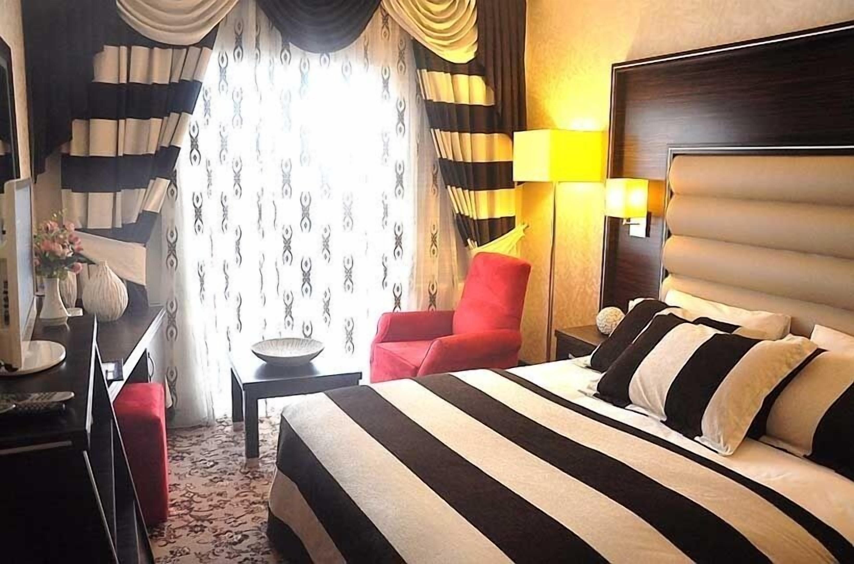 A hotel room with a large bed, a red armchair, a bedside table with a lamp, and curtains with a striped pattern.
