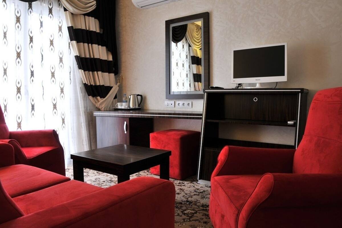 A hotel room with a red sofa set, a small table, a TV, a mirror, and a kitchenette area.