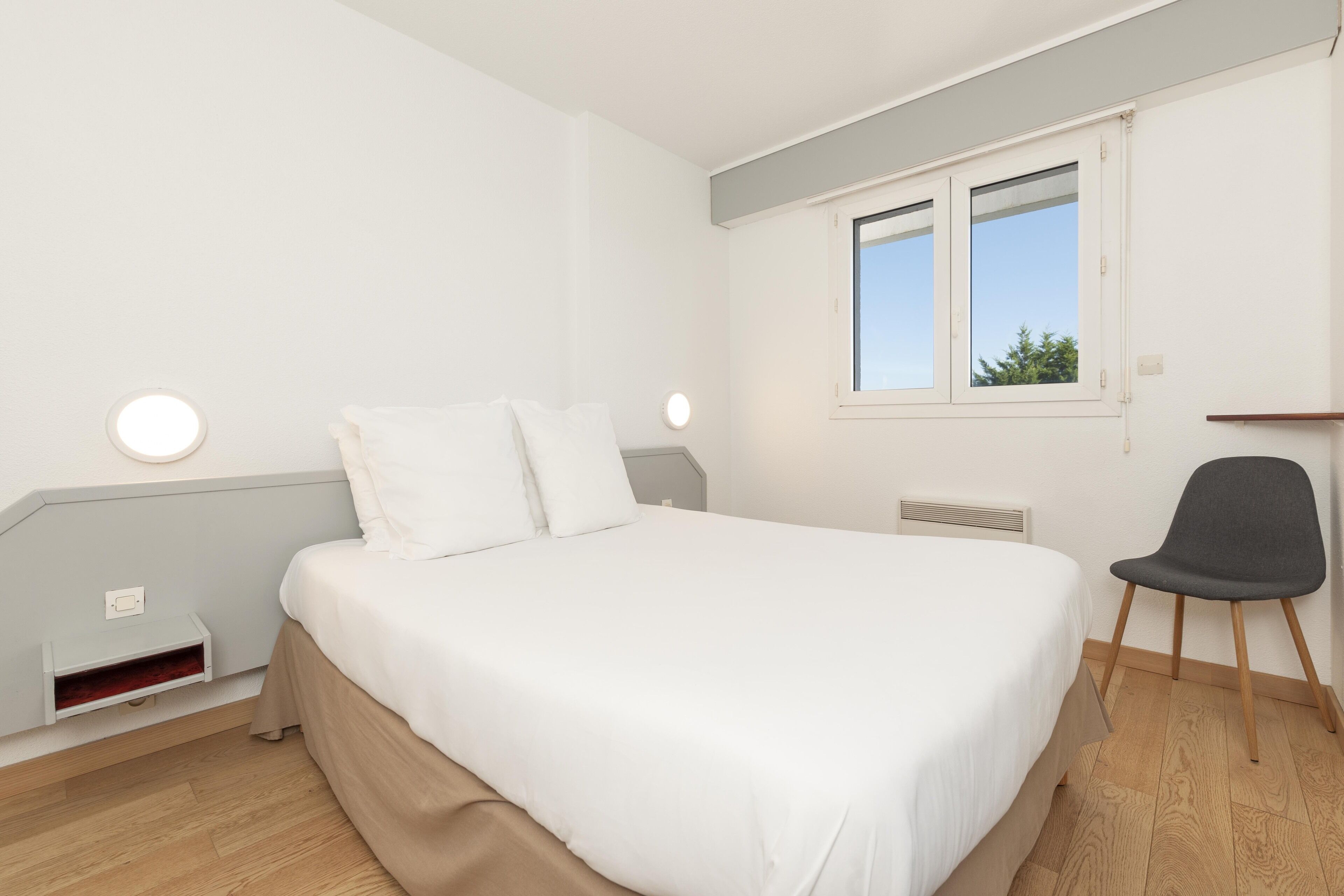 Un dormitorio con cama, una silla, una ventana y una mesita de noche.