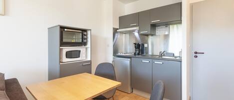 A compact kitchen with a microwave, stove, and refrigerator. There is a wooden table with two chairs and a brown sofa.