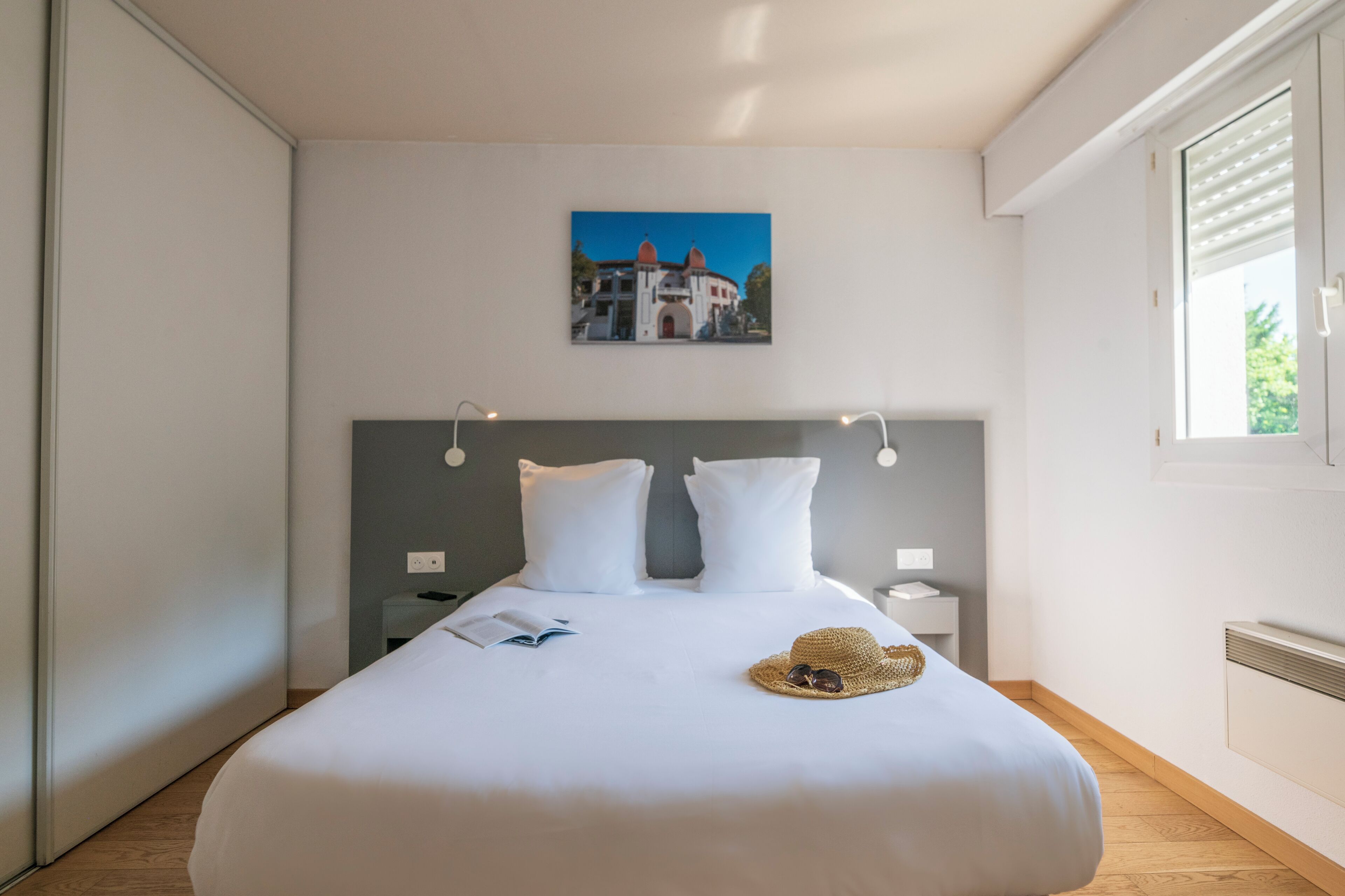 A neatly made bed with white linens, a headboard with wall-mounted lights, a small bedside table with a book and a straw hat, and a framed picture on the wall.