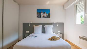A neatly made bed with white linens, a headboard with wall-mounted lights, a small bedside table with a book and a straw hat, and a framed picture on the wall.