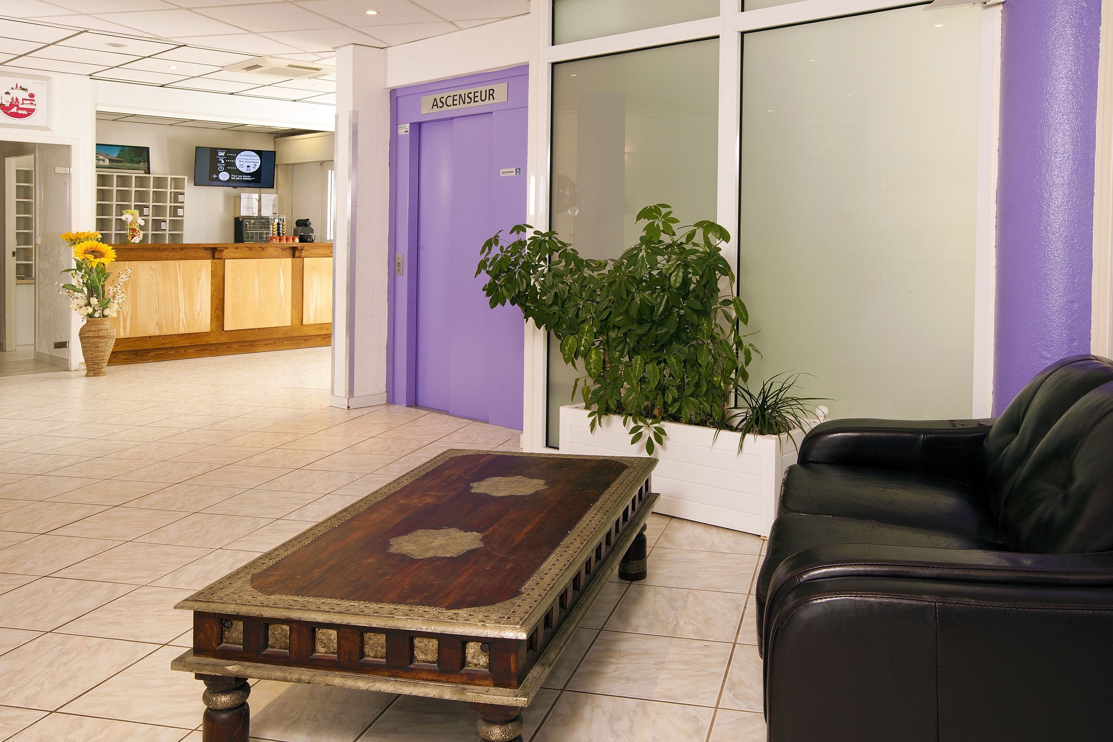 A reception area with a wooden reception desk, a purple door labeled "ASCENSEUR," a black leather sofa, and a potted plant.