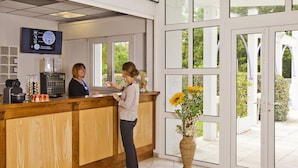 A reception area with a wooden counter, a reception desk, a television mounted on the wall, and a large window with a view of greenery outside. - CERISE Dax - Les Jardins du Lac (Saint-Paul-les-Dax)
