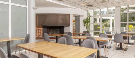 A modern dining area with wooden tables and grey chairs, a large central fireplace, and large windows allowing natural light.