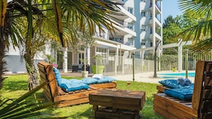 A poolside area with wooden loungers and a wooden table, surrounded by greenery and a building with balconies. - CERISE Dax - Les Jardins du Lac (Saint-Paul-les-Dax)