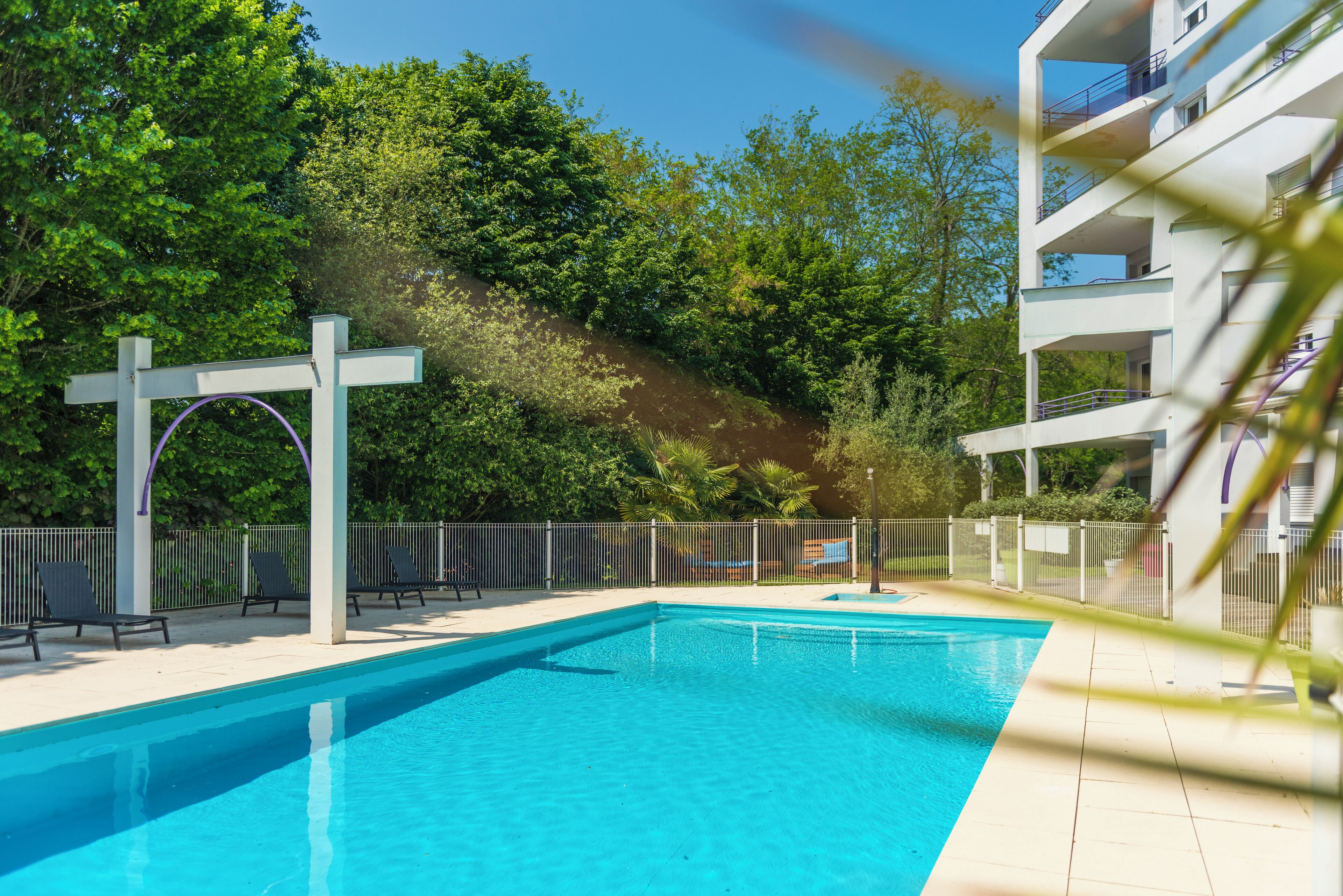 A swimming pool with a white pergola, surrounded by a fence, and a building with balconies in the background.