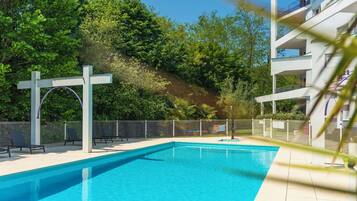 A swimming pool with a white pergola, surrounded by a fence, and a building with balconies in the background.