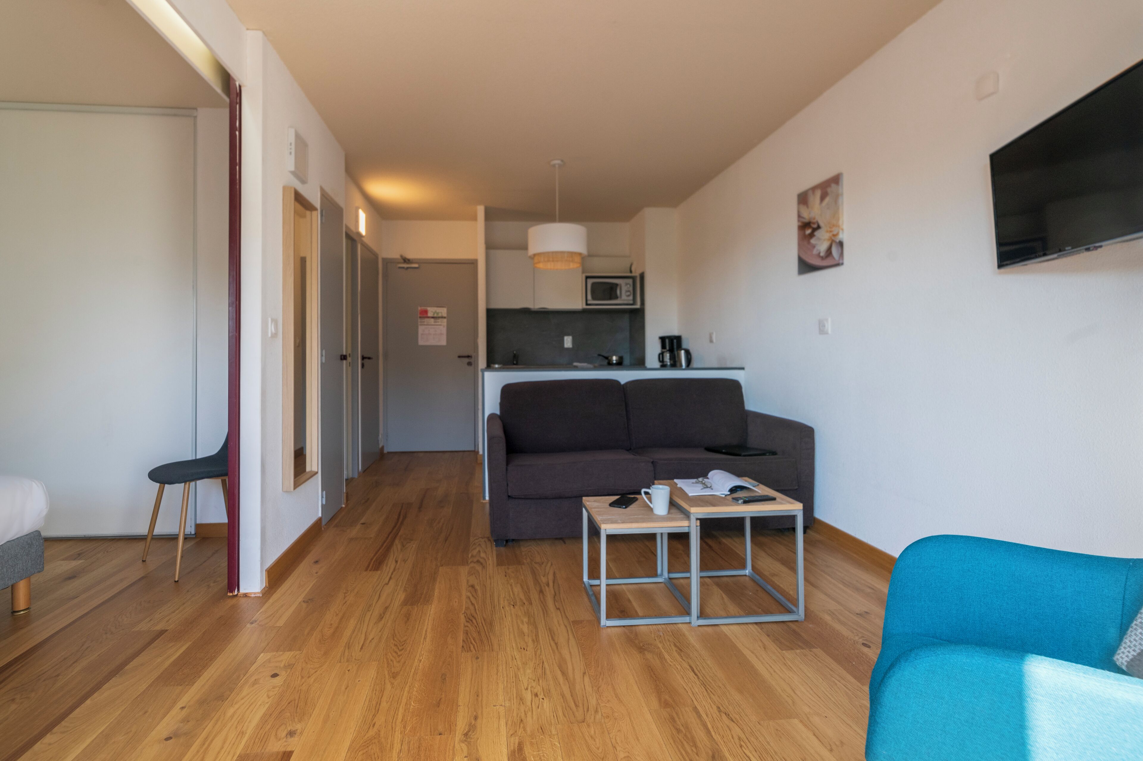 Een moderne woonkamer met een bank, een salontafel en een televisie. Er is een keukenhoek met een magnetron en een hanglamp.