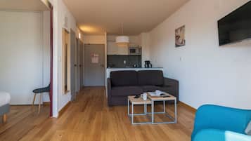 Een moderne woonkamer met een bank, een salontafel en een televisie. Er is een keukenhoek met een magnetron en een hanglamp.