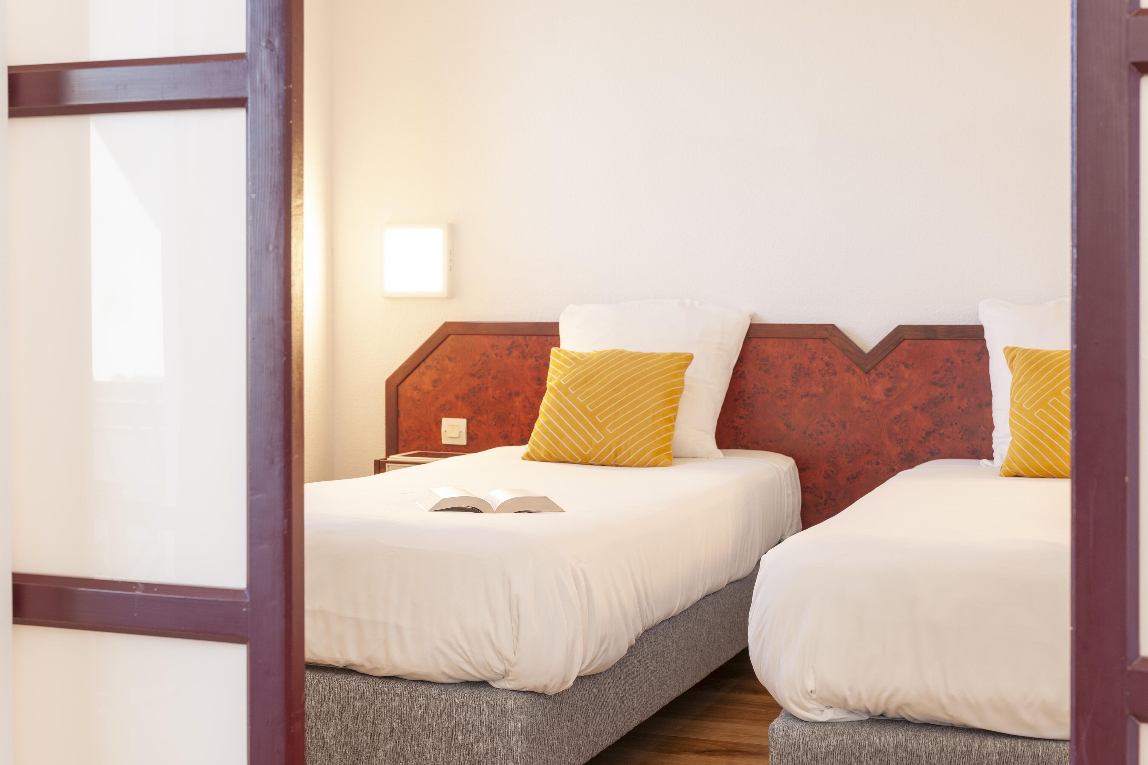 Ein Hotelzimmer mit zwei Betten, jeweils mit weißer Bettwäsche und einem hölzernen Kopfteil. An einem Bett befindet sich ein Nachttisch mit einer Lampe und einem offenen Buch.