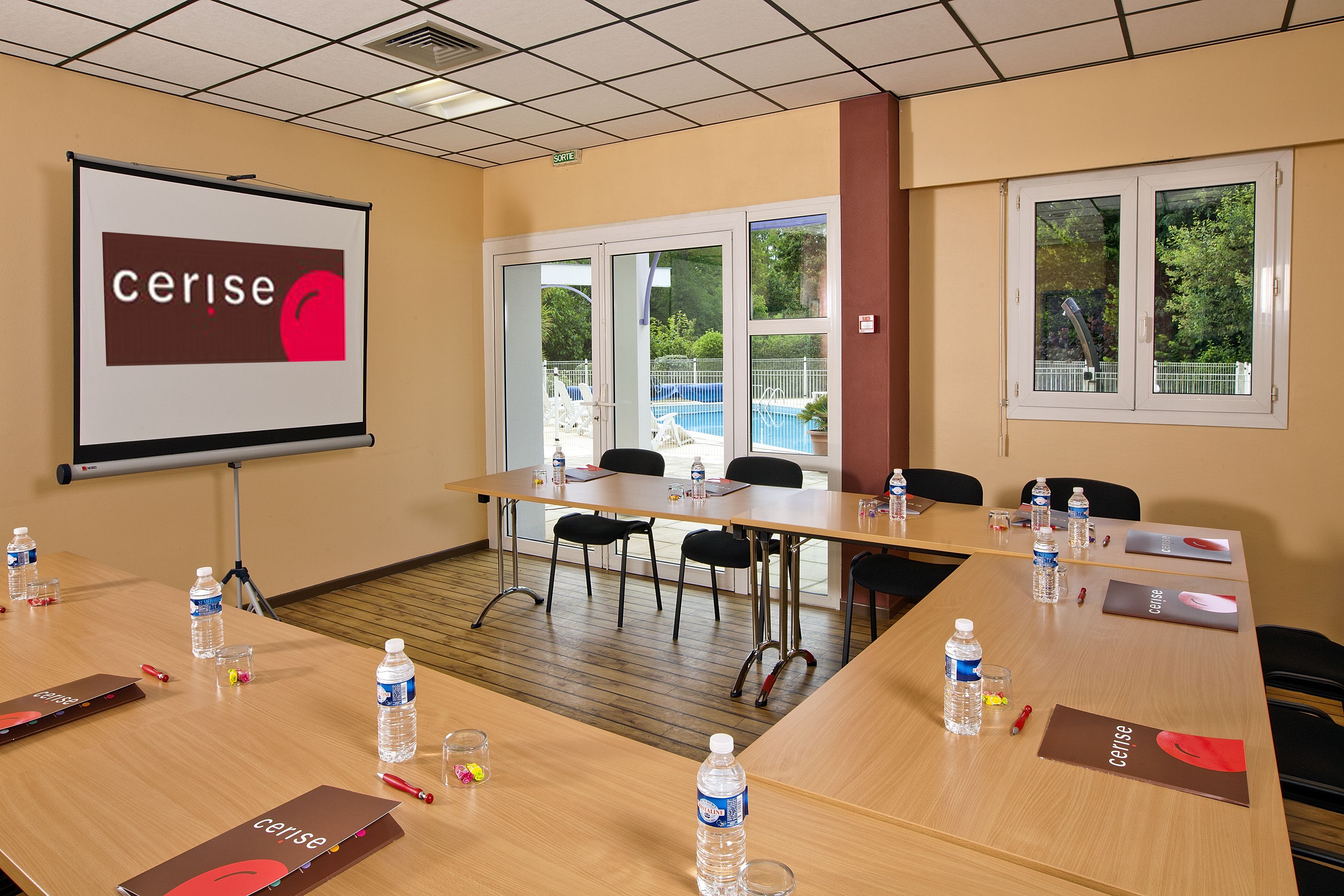 Un salón de conferencias con una pantalla grande mostrando 'cerise', una mesa de madera con botellas de agua, tarjetas de identificación y un proyector.