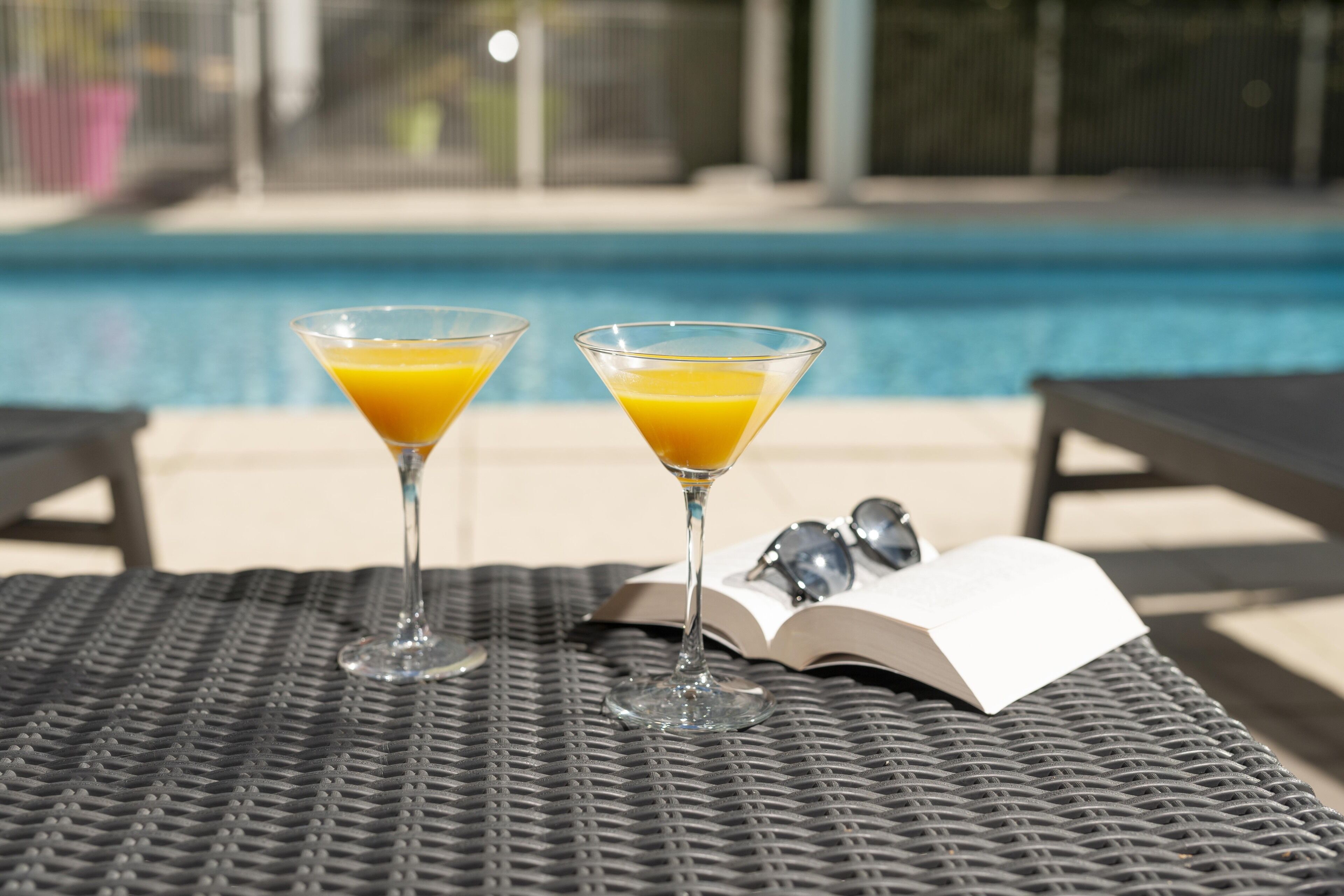Two martini glasses with orange juice on a woven table by a poolside.