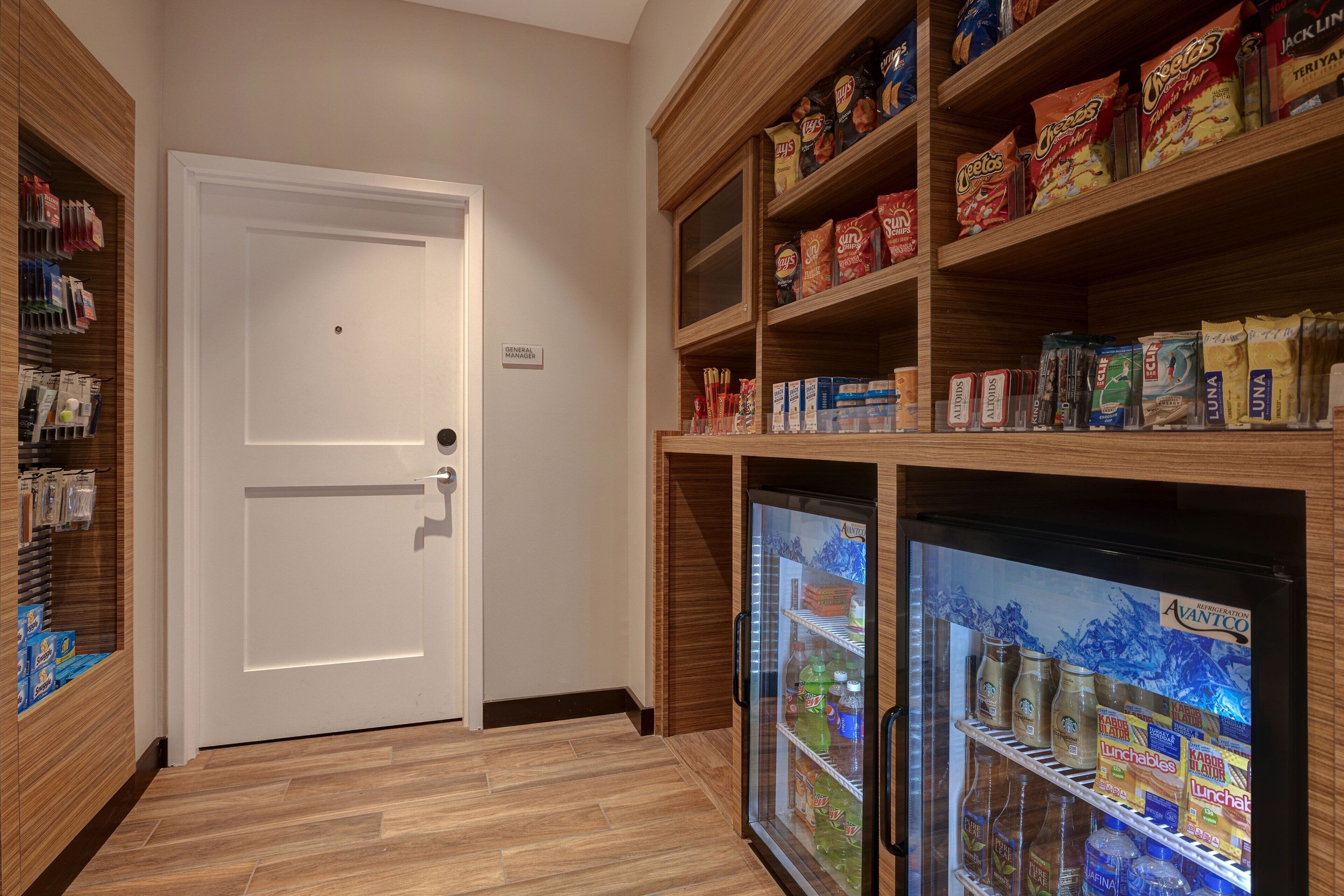 A hotel pantry with shelves stocked with snacks and beverages, a refrigerator, and a door labeled "General Manager."
