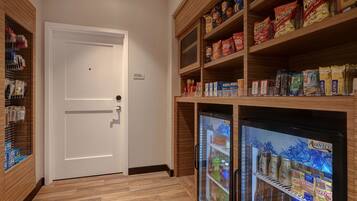 Una despensa de hotel con estantes llenos de refrigerios y bebidas, un refrigerador y una puerta con la etiqueta 'Gerente General'.