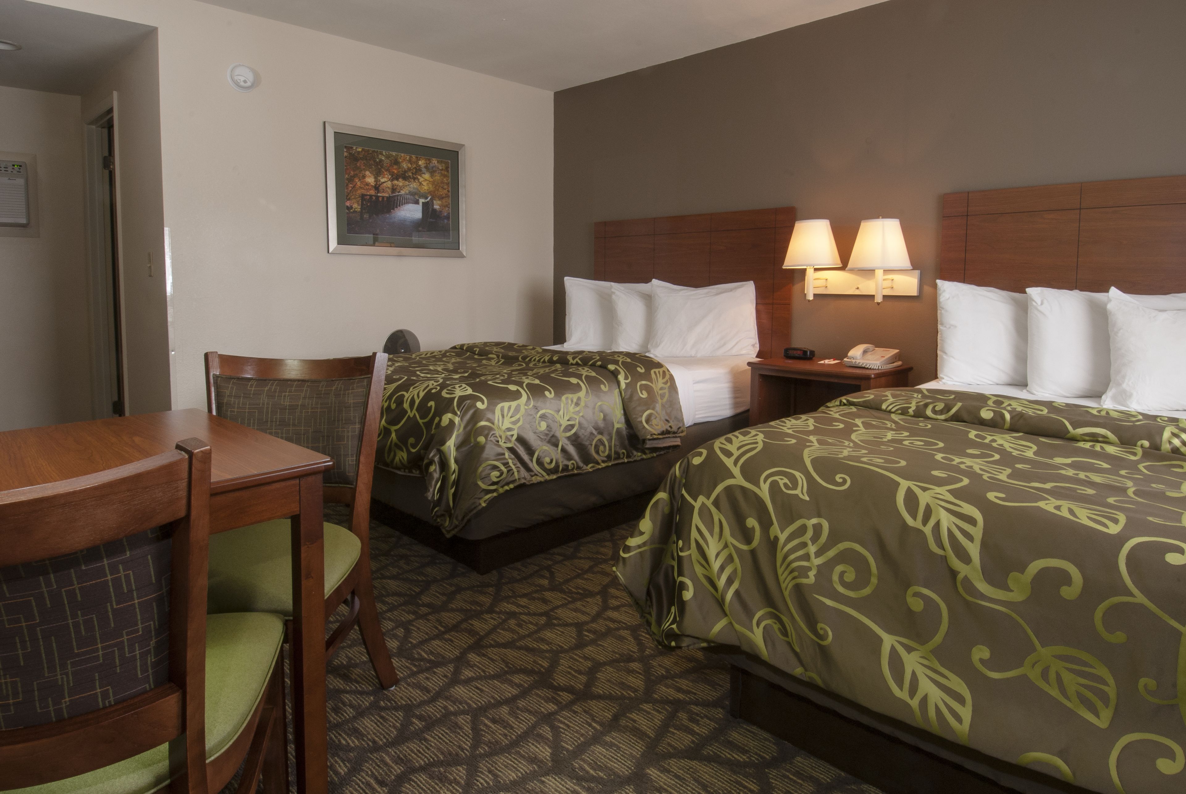 A hotel room with two beds, a desk, and a chair. There is a painting on the wall and two bedside lamps.