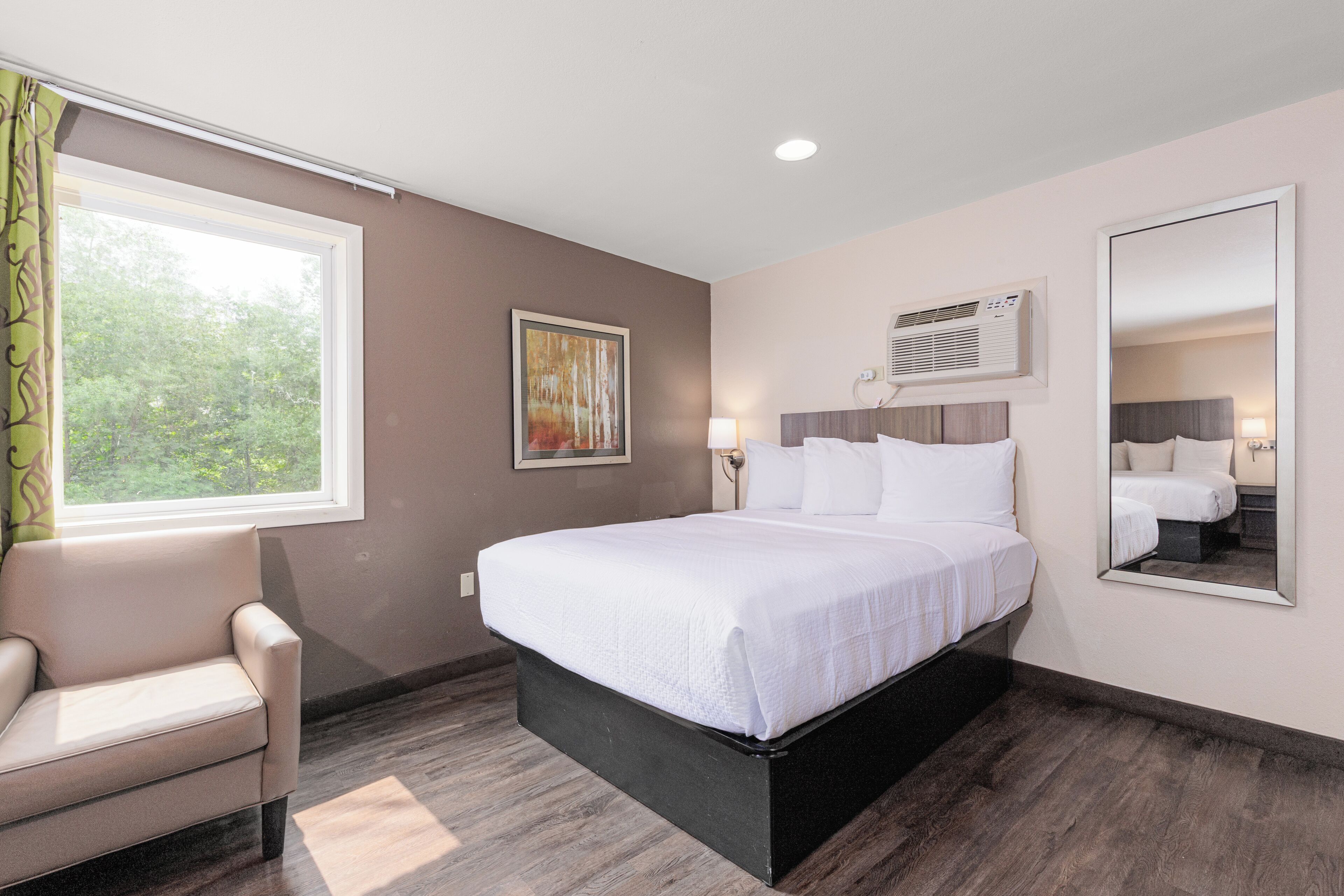 Ein Hotelzimmer mit einem großen Bett, einem Sessel, einem Fenster mit Vorhängen, einem Gemälde und einer Klimaanlage.