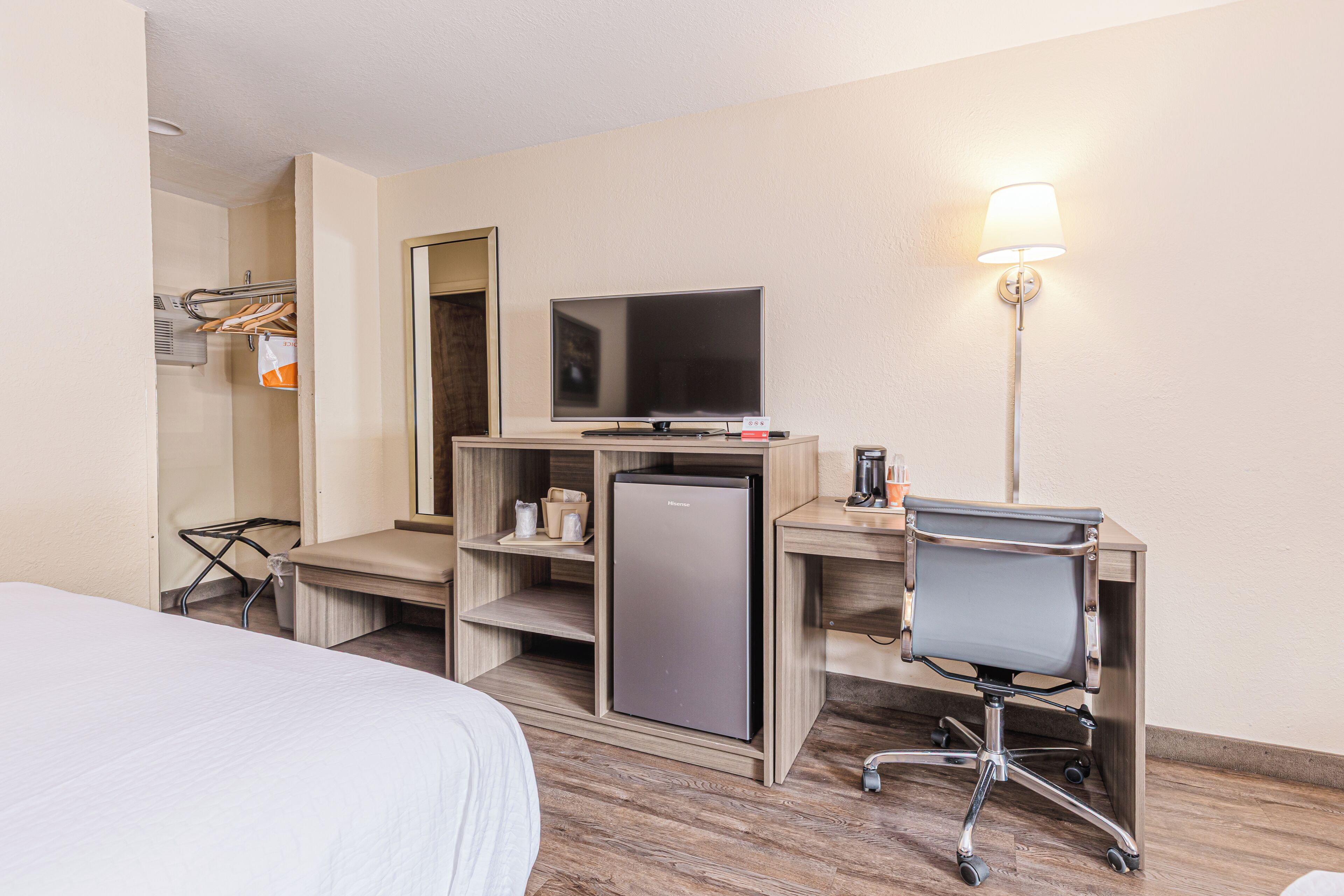Una habitación de hotel con una cama, un escritorio con computadora, un refrigerador, un televisor y una lámpara.