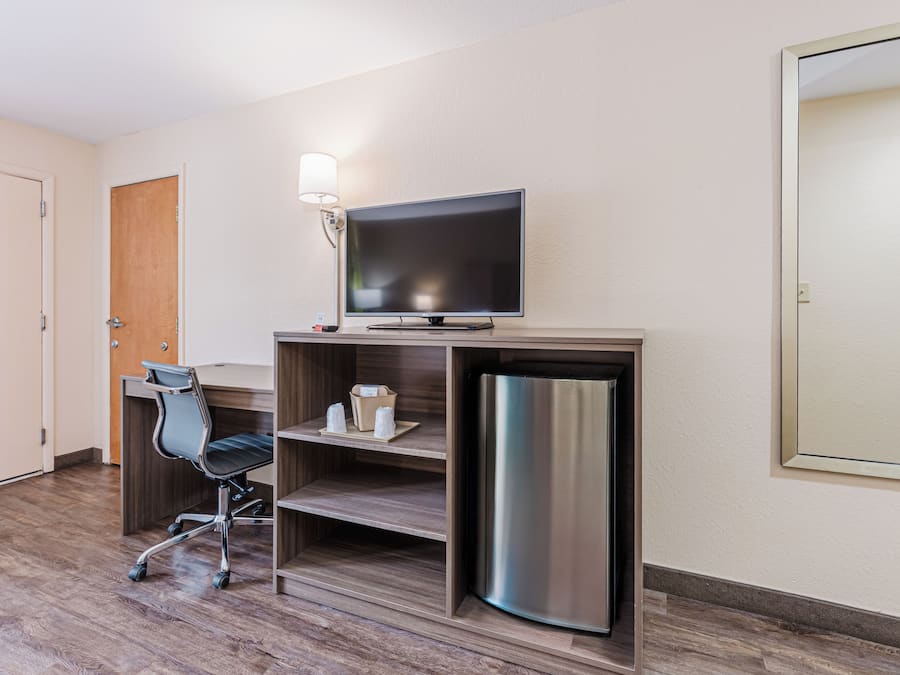 A hotel room with a wooden desk, a chair, a flat-screen TV, a mini-fridge, and a lamp.