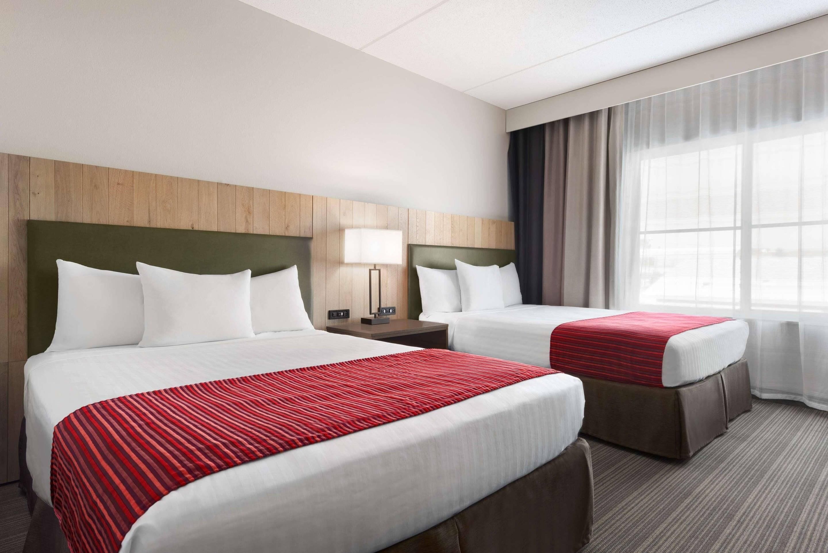 A hotel room with two beds, a wooden headboard, a nightstand with a lamp, and a window with curtains.