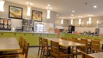 Un restaurante con armarios verdes, mesas y sillas de madera. Se observan dispensadores de comida, una máquina de café y obras de arte en las paredes.