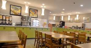 A restaurant with green cabinets, wooden tables, and chairs. There are food dispensers, a coffee machine, and artwork on the walls.