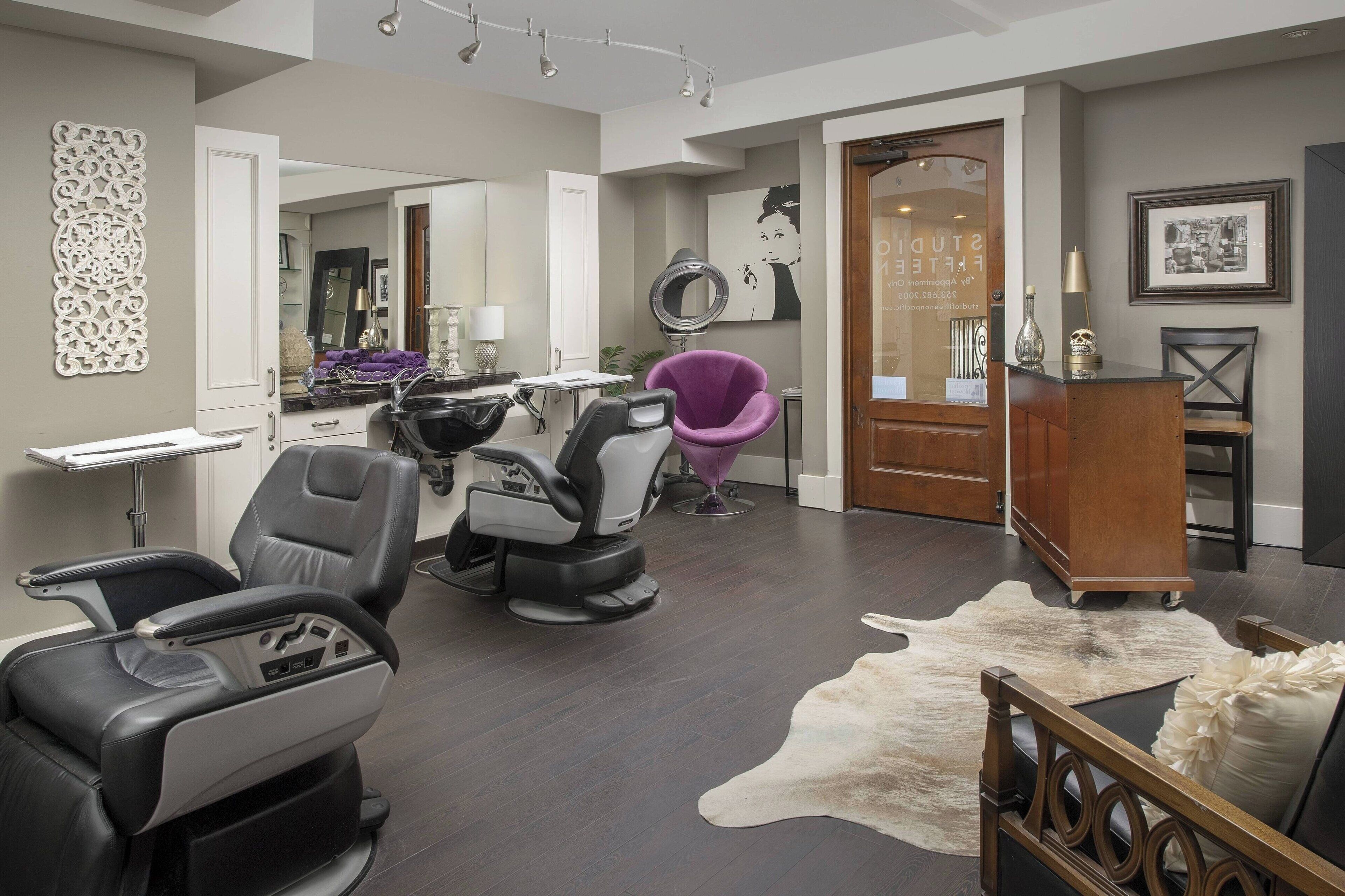 A modern hair salon with multiple seating areas, including a large salon chair, a purple chair, and a wooden bench. There is a large mirror, a hairdryer, and a sink. The room has a dark wooden floor and a cowhide rug.