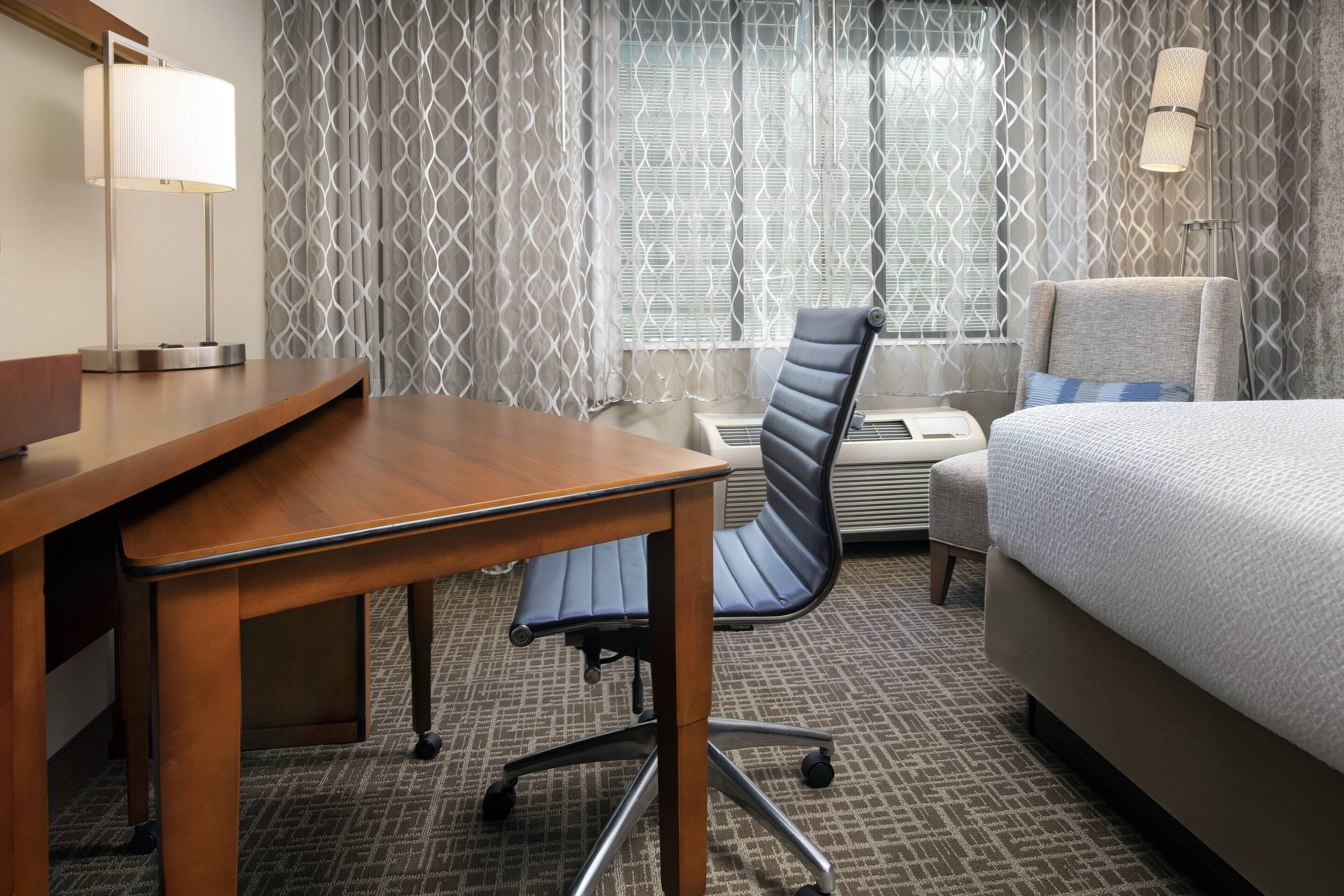 Una habitación de hotel con un escritorio de madera, una silla moderna, una cama y una cortina estampada.