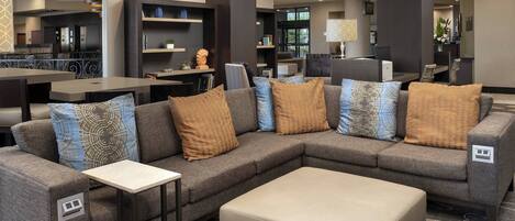 A modern hotel lobby with a sectional sofa, a small table, and a large ottoman. There are multiple seating areas and a reception desk in the background.