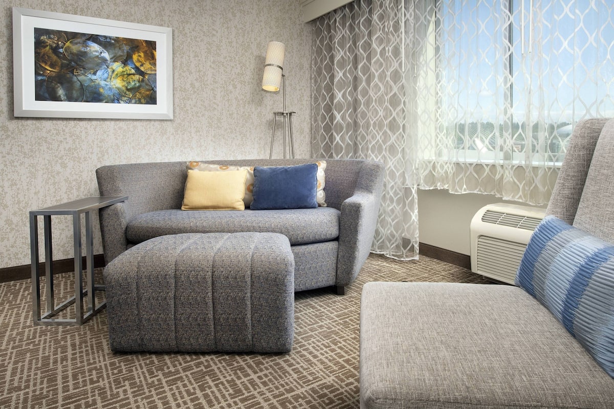 Uma sala de estar moderna com um sofá cinzento, um banquinho e uma poltrona menor. Há um abajur, uma obra de arte emoldurada na parede e uma janela com vista.