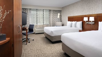 A hotel room with two beds, a desk with a chair, a TV, and a window with patterned curtains.