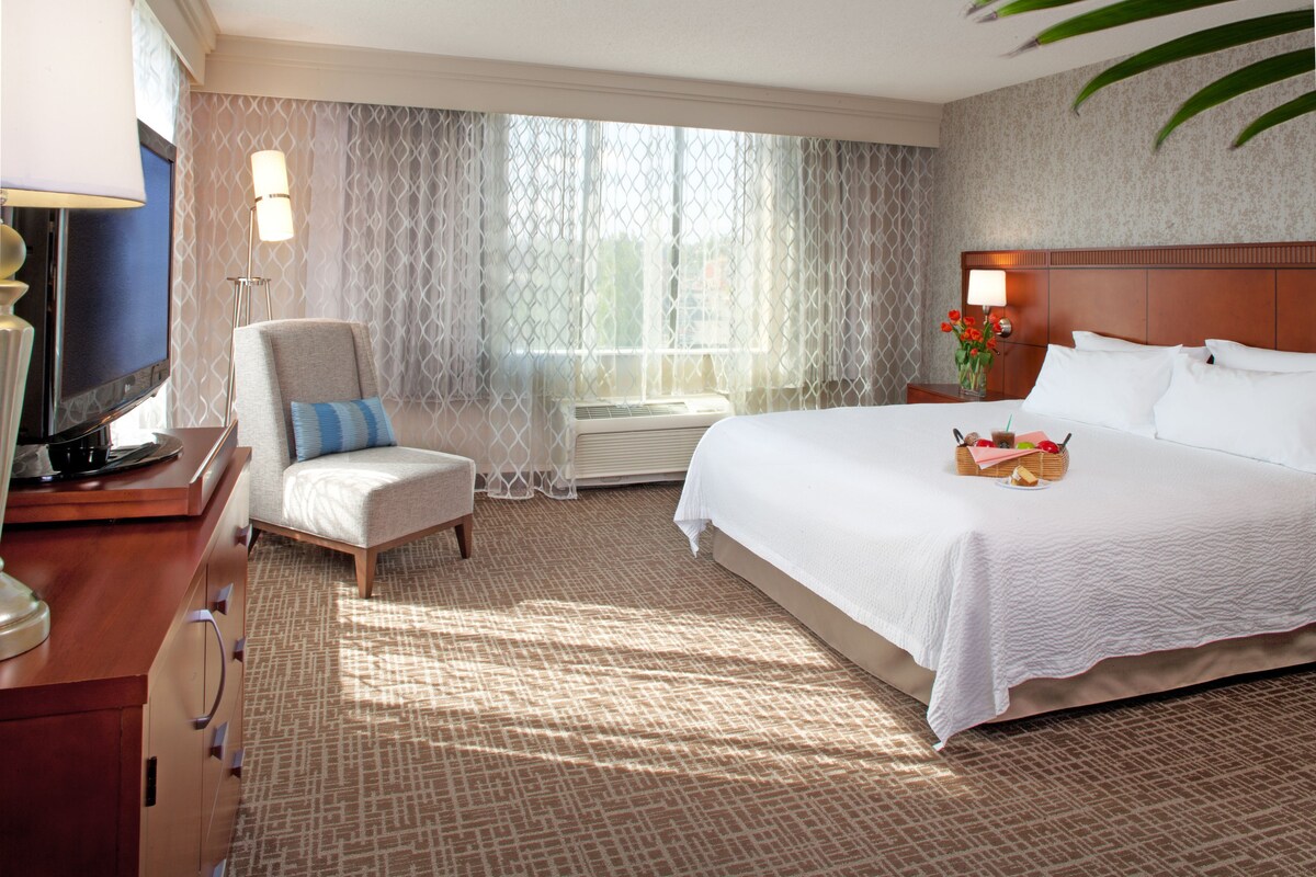 A hotel room with a large bed, a chair, a desk with a lamp, and a window with curtains.
