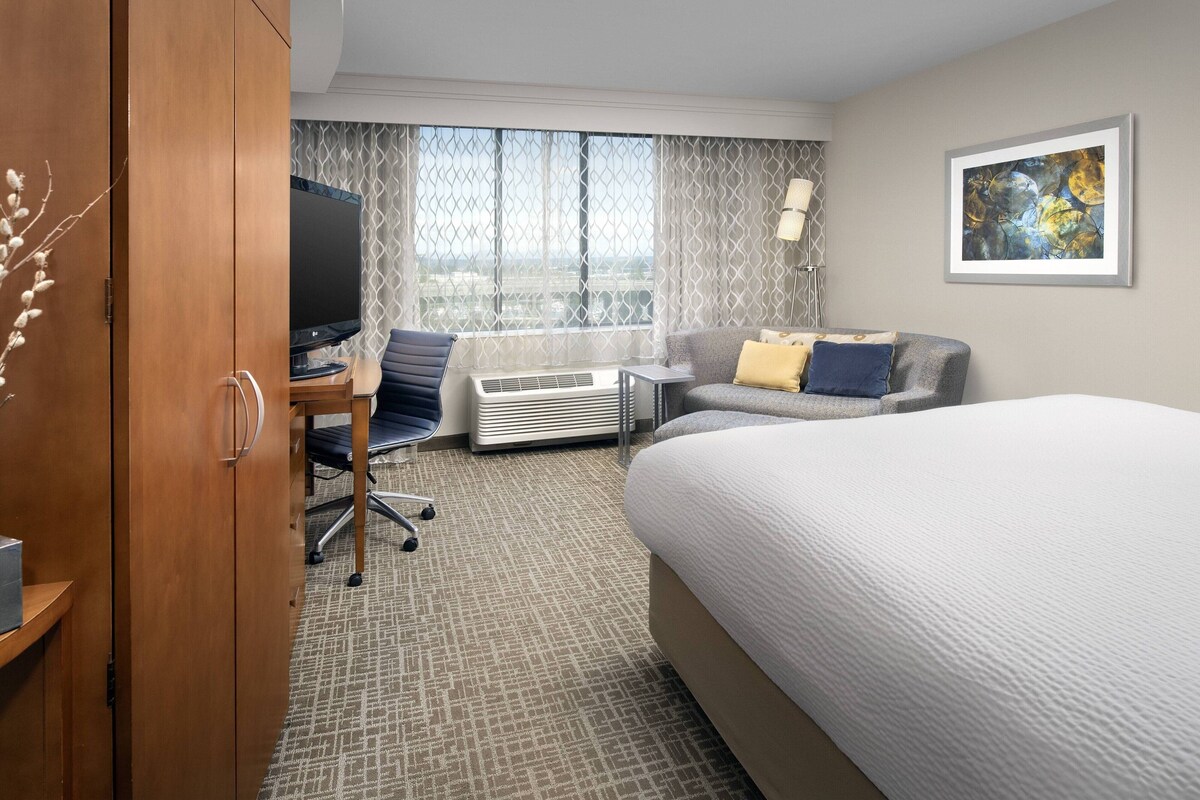 A hotel room with a large bed, a desk with a computer, a chair, a sofa with cushions, a TV, and a window with curtains.